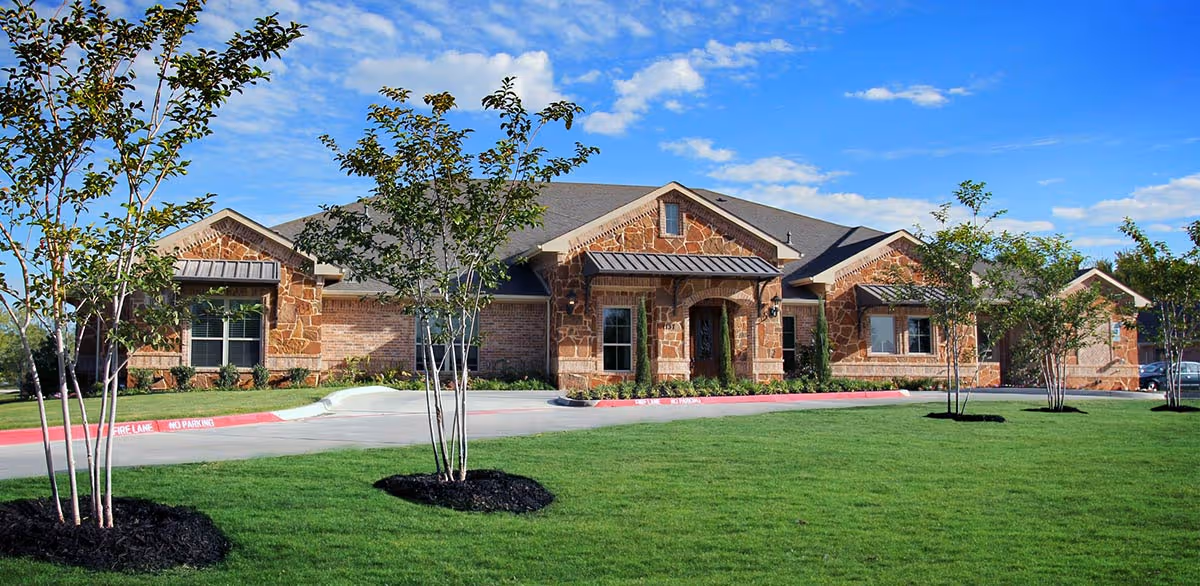 Exterior view of a single-story building with stone and brick facade, surrounded by a well-maintained lawn and young trees under a blue sky with scattered clouds.