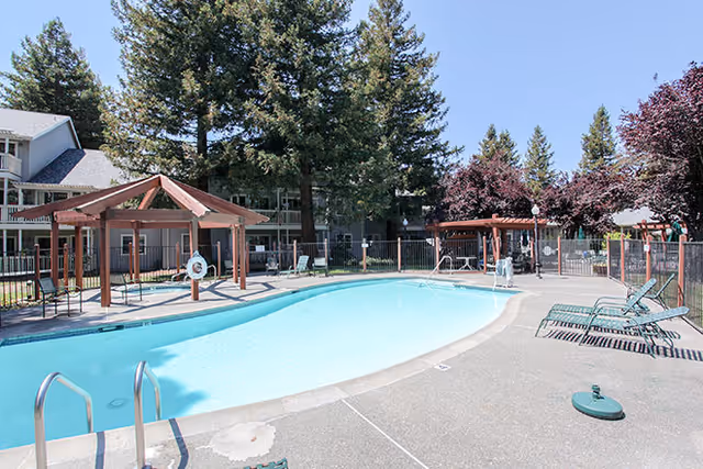 Outdoor swimming pool with lounge chairs, wooden pergolas, and surrounding apartment buildings and trees.