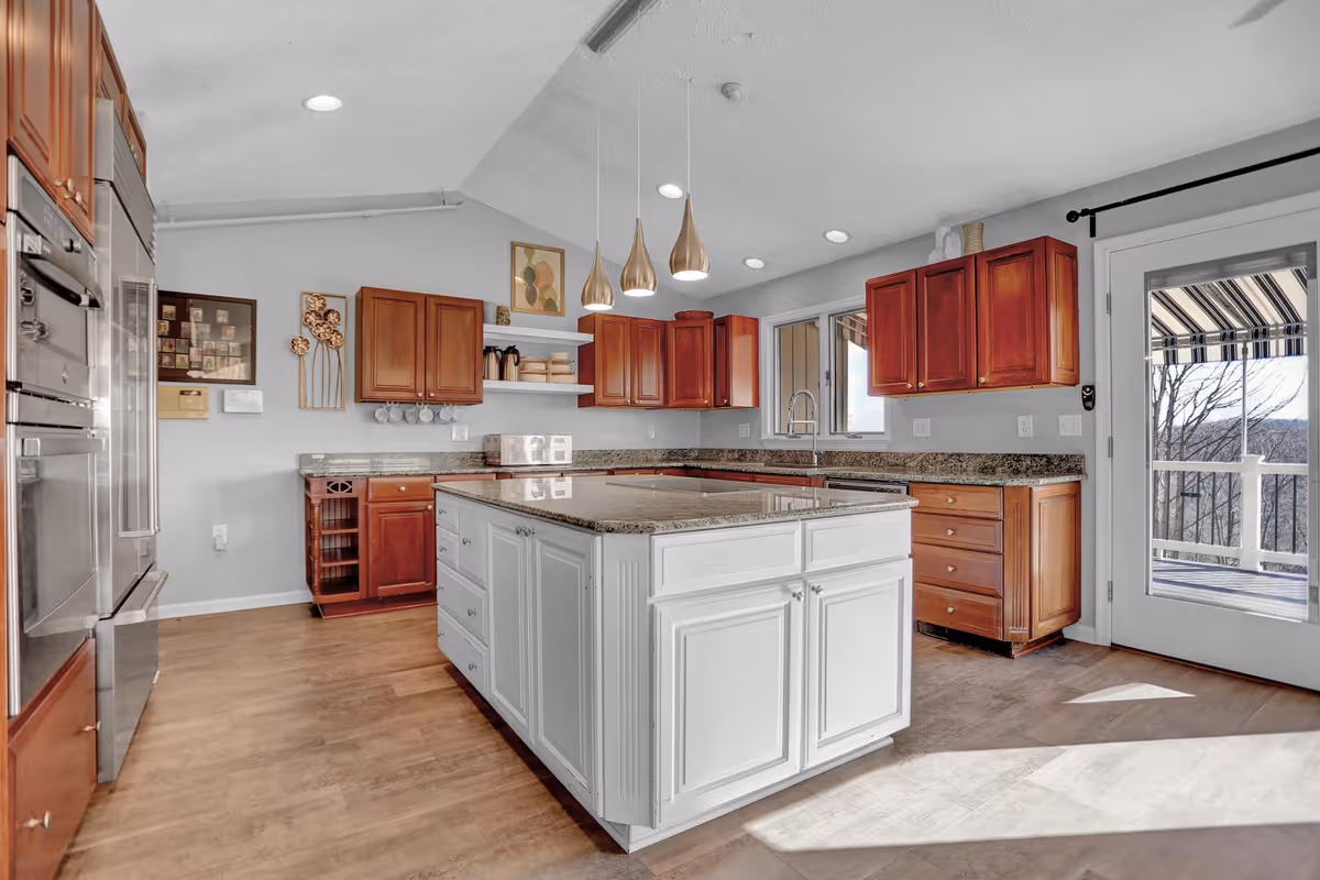 Bright kitchen with wooden cabinets and a large white island in the center topped with granite countertops. Three pendant lights hang above the island. Stainless steel appliances are built into the cabinetry on the left. A door with glass panels leads to an outdoor balcony with a striped awning. The kitchen has wooden flooring and light gray walls with some decorative artwork and shelves.