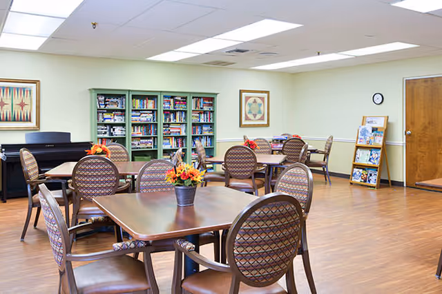 A well-lit common room with several wooden tables and cushioned chairs arranged neatly. Each table has a small flower arrangement. In the background, there is a green bookshelf filled with books and board games, a piano, framed artwork on the walls, a clock, and a wooden door. The floor is wooden and the ceiling has recessed lighting.