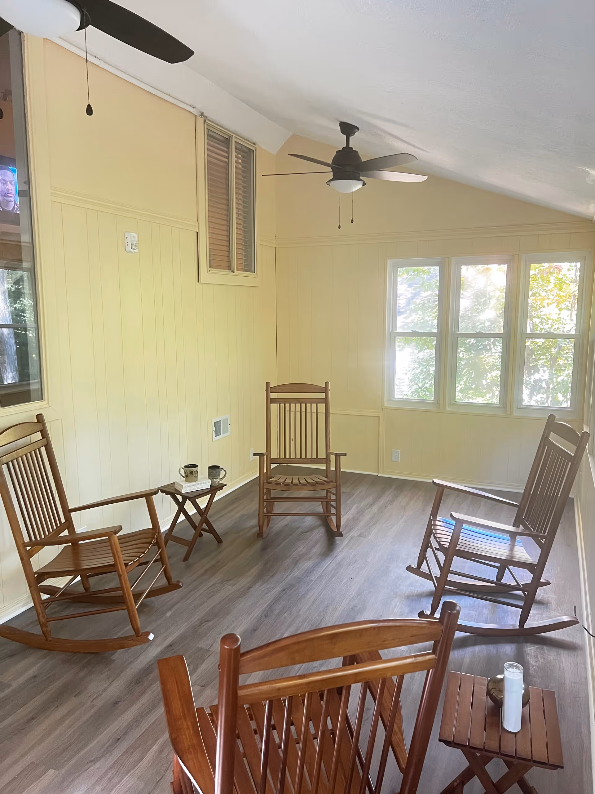 A bright room with four wooden rocking chairs arranged in a circle on a wood floor. There are two small wooden side tables, one with two coffee mugs and a book, and the other with a candle. The walls are painted light yellow, and there are three windows letting in natural light. Two ceiling fans with lights are mounted on the ceiling.