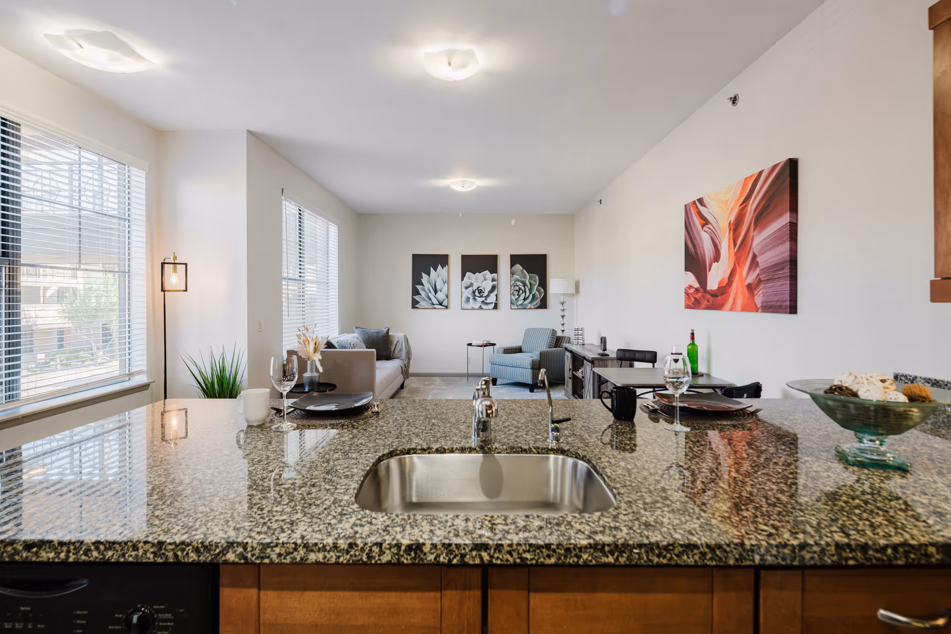 View from a kitchen island with a granite countertop and a built-in sink looking out into a living room area. The living room features a sofa, an armchair, a side table, and wall art including three black and white succulent prints and a colorful abstract painting. Large windows with blinds allow natural light to fill the space.