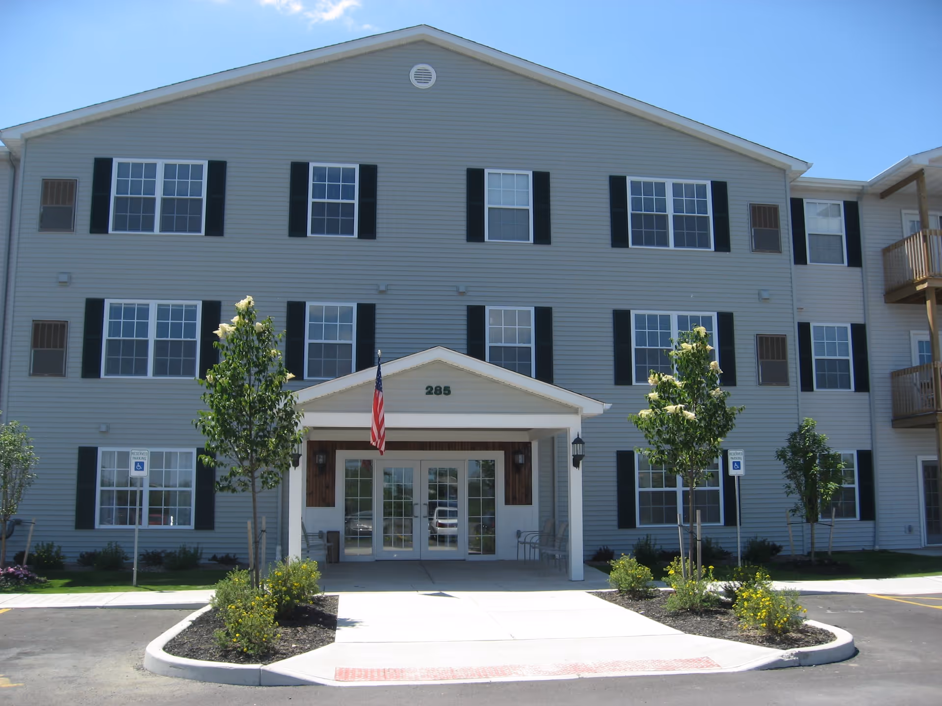 Front exterior view of a three-story senior apartment building with gray siding and black shutters. The entrance has a covered porch with the number 285 above it, an American flag hanging, and landscaped greenery with small trees and bushes along the walkway leading to the door.