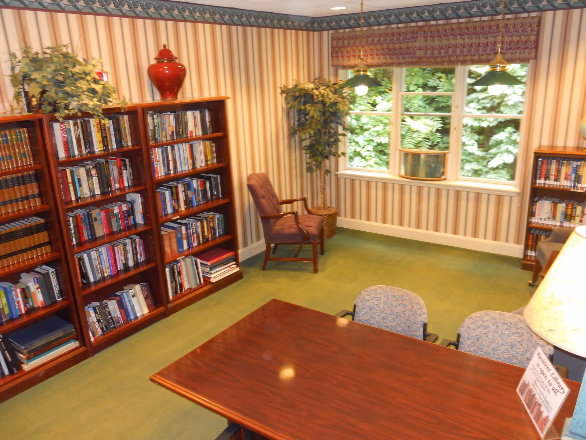 A cozy library room with wooden bookshelves filled with books, a wooden table with two patterned chairs, a purple armchair near a window with a view of green foliage, and a potted plant in the corner. The room has striped wallpaper and green carpet.