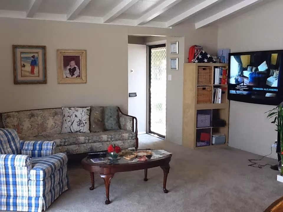 Cozy living room with a floral sofa, plaid armchair, wooden coffee table, wall-mounted TV, and shelving by an open doorway.
