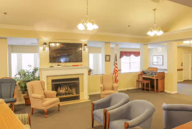 A cozy living room area in a senior living facility with multiple upholstered chairs arranged around a fireplace with a mounted flat-screen TV above it. There is a piano against the far wall near a window with red curtains and an American flag beside it. The room is softly lit with ceiling lights and wall sconces.