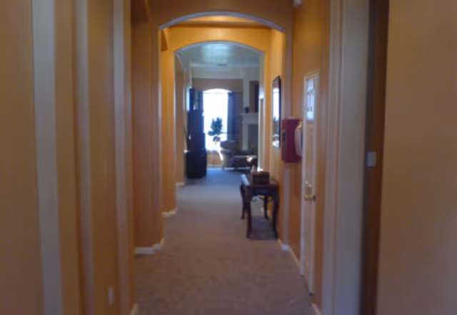 View down a carpeted hallway with beige walls and an arched doorway leading to a living room area with a window, curtains, and furniture including a chair and a table with decorative items.