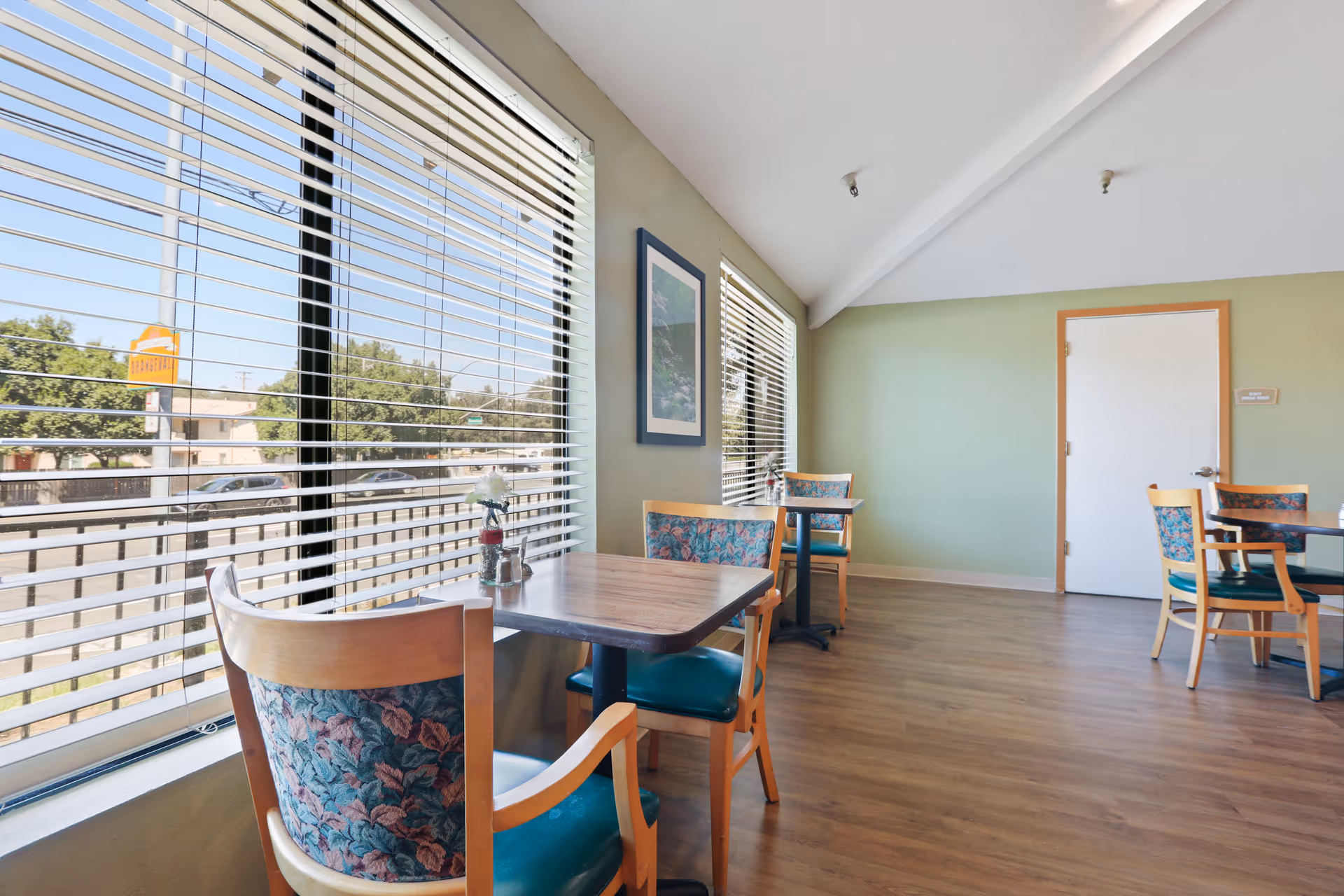 A bright dining area with wooden tables and chairs featuring floral upholstery. Large windows with horizontal blinds allow natural light to fill the room. The walls are painted light green, and there is a framed picture hanging on one wall. A white door with a sign labeled 'Staff Break Room' is visible at the far end of the room.