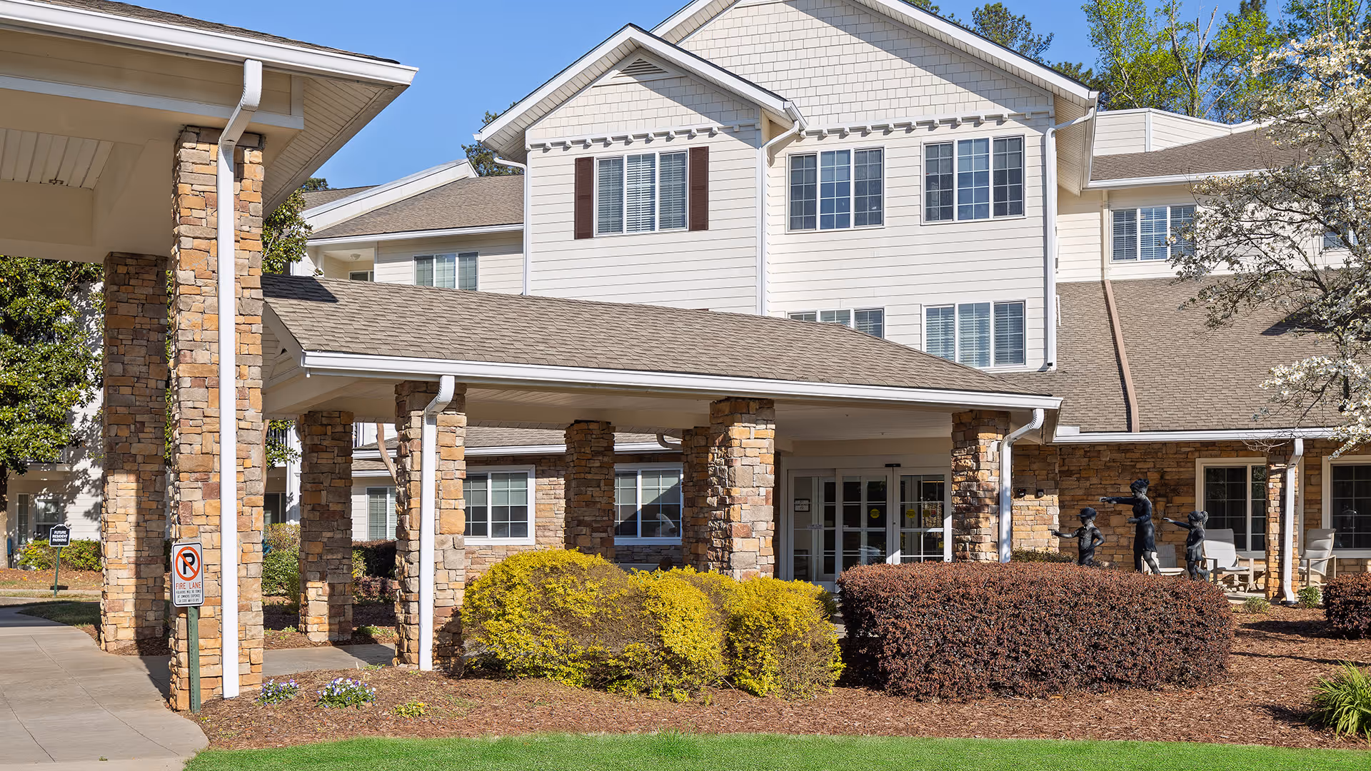 Exterior view of Laurel Grove by Barclay House, showing a multi-story building with white siding and stone pillars supporting a covered entrance. There are bushes and landscaping in front of the building, and a sculpture of children playing near the entrance.