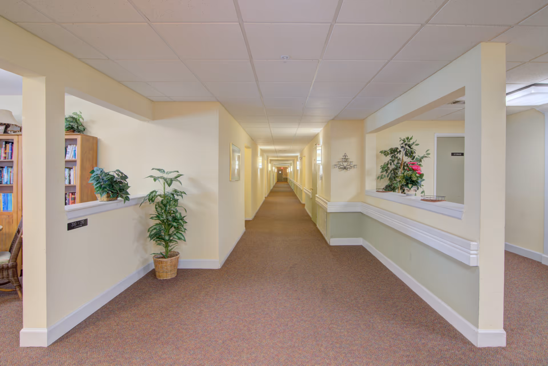 A long, well-lit hallway in a senior living facility with beige walls, carpeted floor, and ceiling tiles. There are plants in pots placed along the walls and a bookshelf visible through an open window-like cutout on the left side. The hallway has handrails on both sides and doors along the corridor.