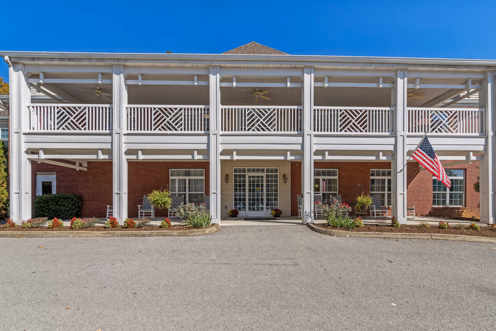 Front exterior view of a two-story brick building with white columns and railings. The entrance has glass double doors with sidelights, flanked by windows and outdoor seating areas with chairs and plants. An American flag is displayed on the right side of the building under a clear blue sky.