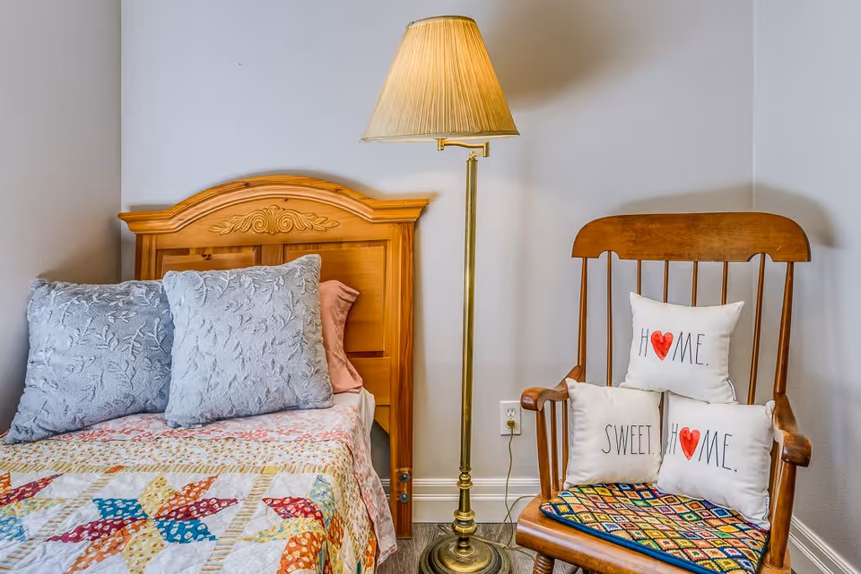 A cozy bedroom corner featuring a wooden bed with a carved headboard, two gray textured pillows, and a colorful patchwork quilt. Next to the bed is a brass floor lamp with a beige lampshade, and a wooden rocking chair with three white pillows that have red heart designs and the words 'HOME.' and 'SWEET.' on them. The chair also has a colorful woven seat cushion. The walls are painted light gray.