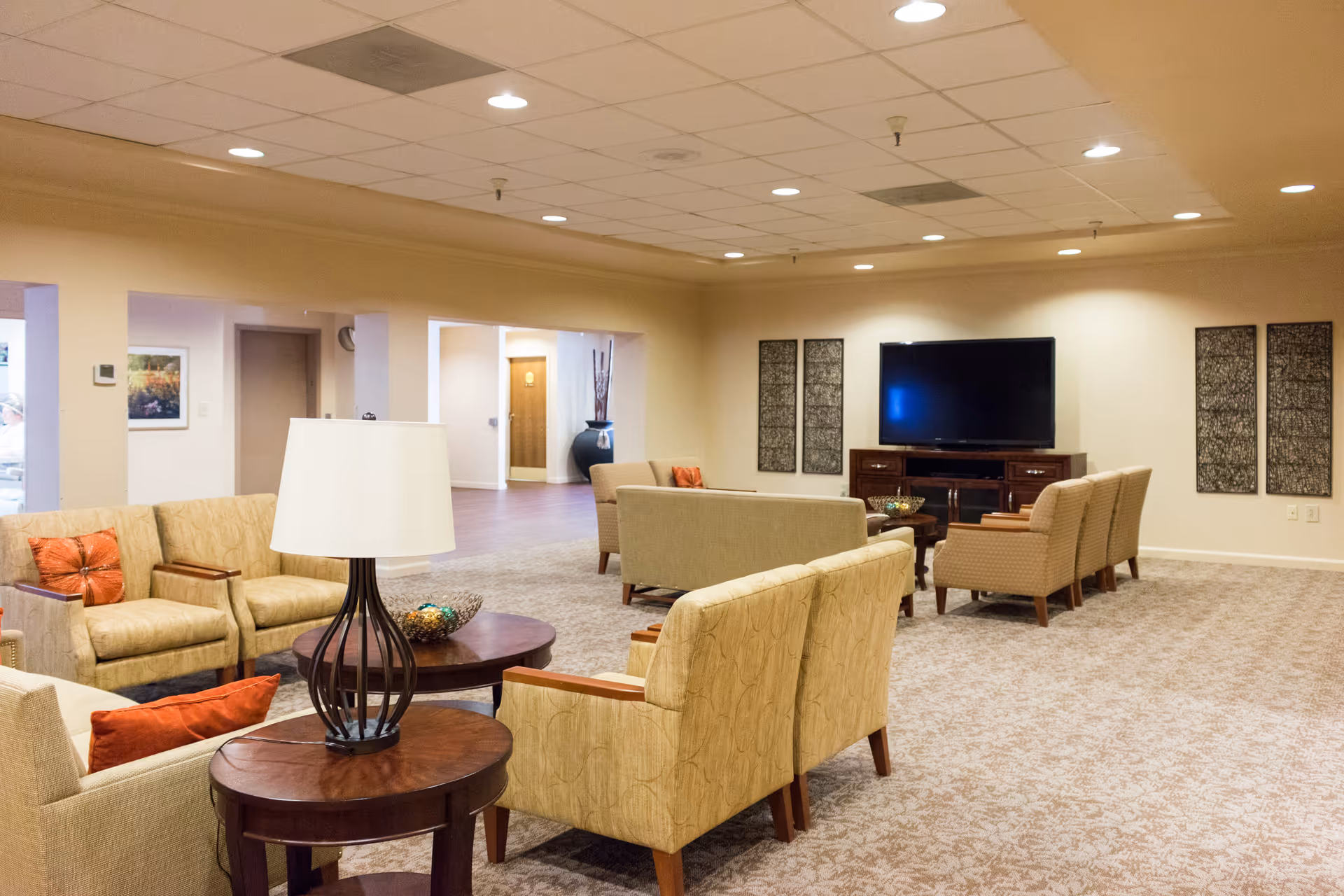 A spacious senior living facility lounge area with beige patterned carpet and cream-colored walls. The room is furnished with multiple beige upholstered chairs and sofas arranged around dark wooden coffee tables. A large flat-screen TV is mounted on a wooden cabinet against the far wall, flanked by decorative wall panels. The ceiling has recessed lighting and a drop ceiling design.
