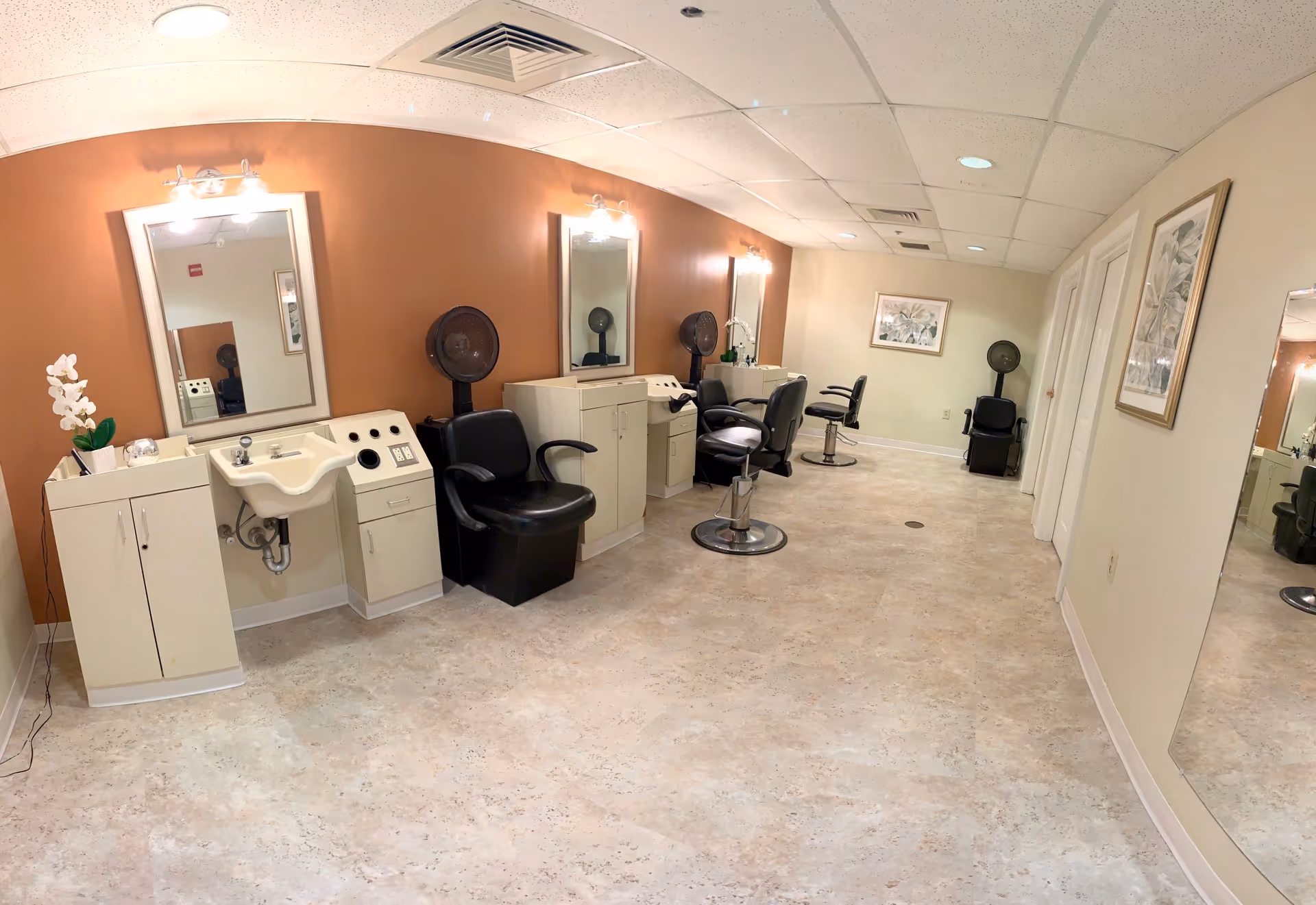 Interior view of a salon area in a senior living facility with three styling stations, each equipped with a mirror, a black salon chair, and a hair dryer. The walls are painted in warm tones with framed artwork, and the floor is covered with light-colored tiles.