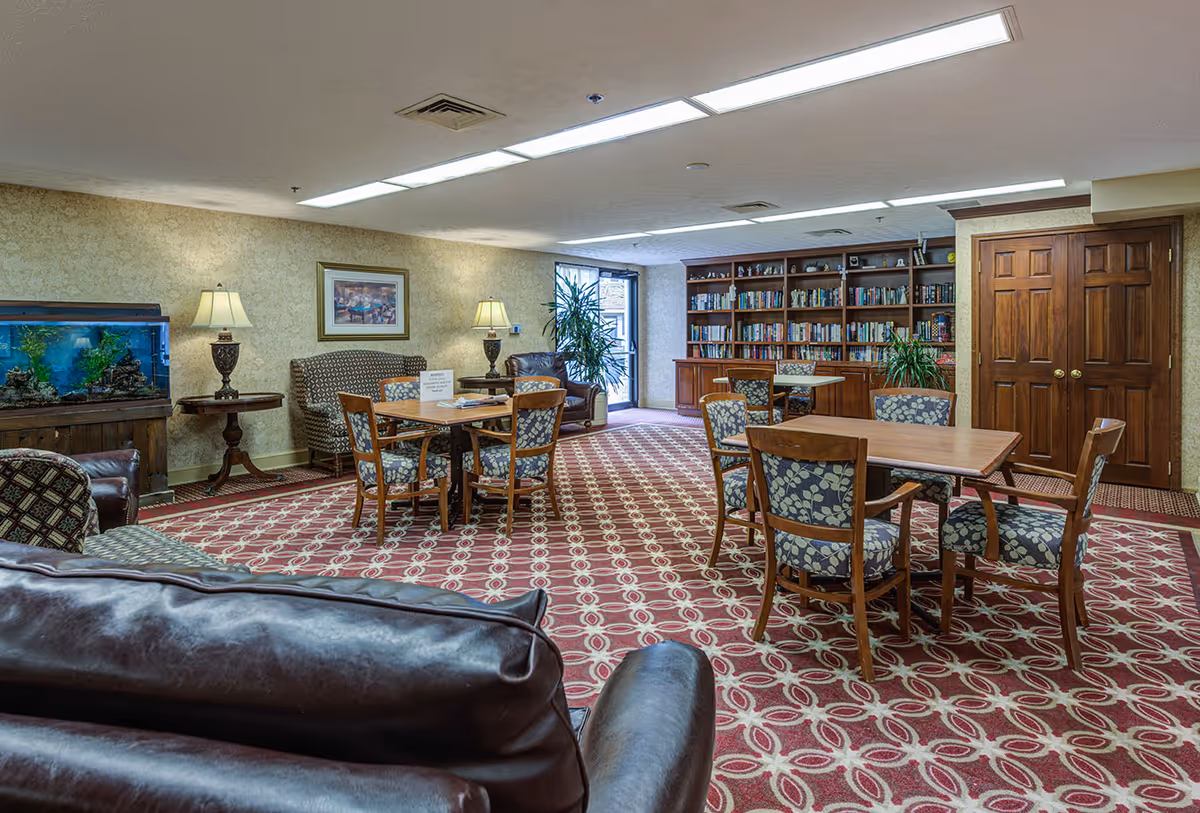 A cozy common area in a senior living facility featuring patterned carpet, several wooden tables with floral upholstered chairs, a large bookshelf filled with books, a fish tank on a wooden stand, two table lamps on side tables, a framed picture on the wall, and a leather couch in the foreground.