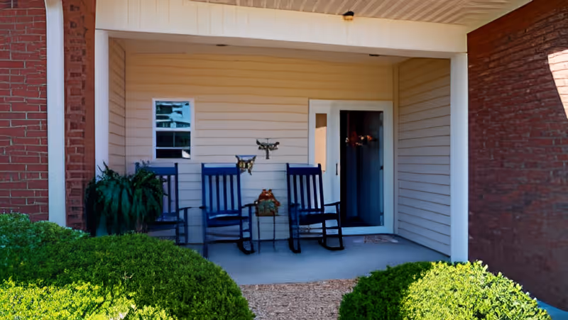 Covered porch area with two blue rocking chairs, a small table with a decorative item, a potted plant, and a door leading inside. The porch is framed by brick walls and beige siding, with green bushes in the foreground.