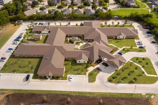 Aerial view of Legend Oaks Healthcare and Rehabilitation facility in Fort Worth, showing a large, single-story building with multiple wings, surrounded by green lawns, parking lots with several cars, and nearby residential houses.