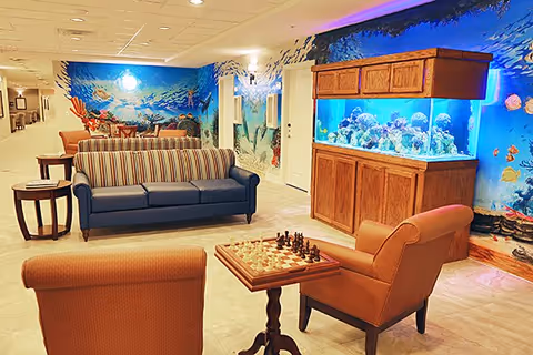 A cozy indoor lounge area featuring a striped sofa, two armchairs, and a wooden chess table. The room has a large aquarium with colorful fish and coral, and the walls are decorated with an underwater ocean mural. The space is well-lit with ceiling lights and has a warm, inviting atmosphere.