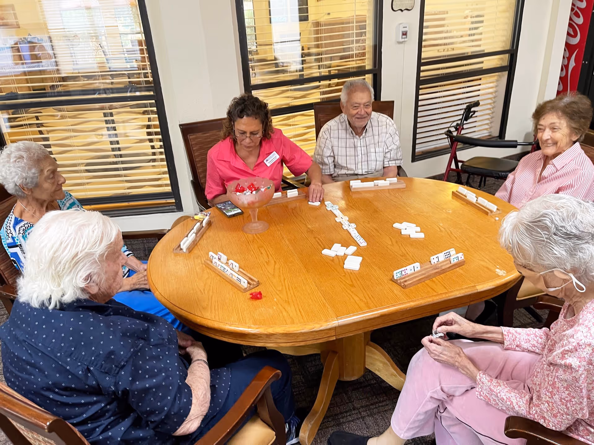 A group of elderly people and a caregiver sitting around a wooden table playing a game of dominoes in a well-lit room with large windows and blinds.