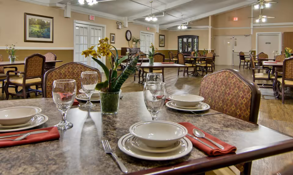Dining room with several set tables, patterned chairs, floral centerpieces, and overhead lighting.