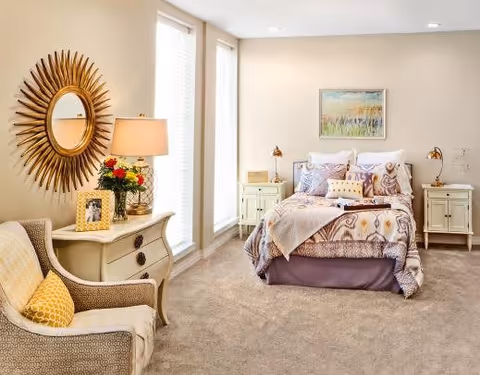 A cozy bedroom with a neatly made bed featuring patterned bedding and multiple pillows. On either side of the bed are matching nightstands with lamps. A framed painting hangs above the bed. To the left, there is a cushioned armchair with a yellow pillow, and a small dresser with a table lamp, a vase of flowers, and a framed photo. A decorative sunburst mirror hangs on the wall above the dresser. The room has beige walls and carpeted floor with tall windows letting in natural light.
