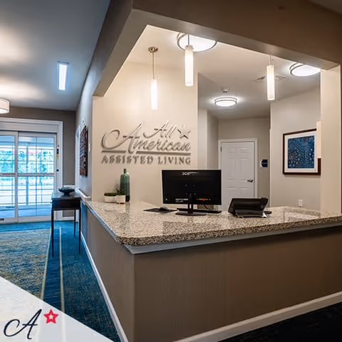 Reception desk area inside All American Assisted Living facility with a granite countertop, computer monitor, phone, and decorative items. The wall behind the desk displays the facility's name. There are pendant lights hanging from the ceiling and a hallway leading to a door. A glass door and windows are visible in the background.