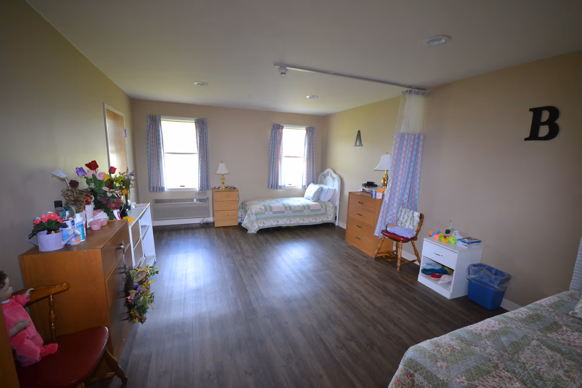 A spacious assisted living bedroom with two single beds, wooden dressers, chairs, and windows with curtains. The room has wood flooring and beige walls with letters 'A' and 'B' on the walls above the beds. Various personal items and decorations are placed on the furniture.