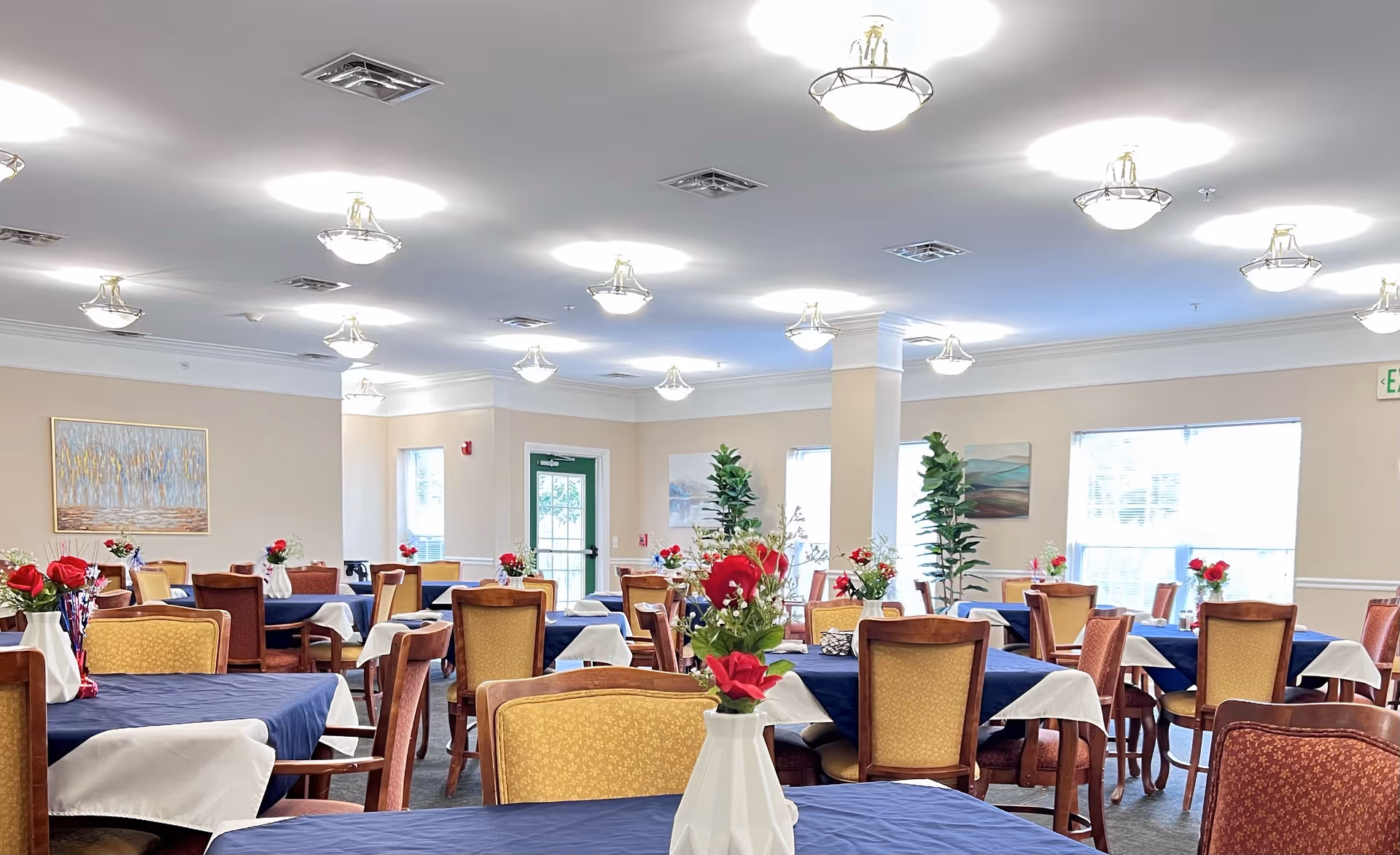 A bright dining room with multiple tables covered in blue tablecloths and white overlays, each adorned with a vase of red and white flowers. The room has beige walls, several windows letting in natural light, and ceiling lights evenly spaced. Chairs with wooden frames and upholstered seats surround the tables. There are paintings and tall green plants decorating the room.