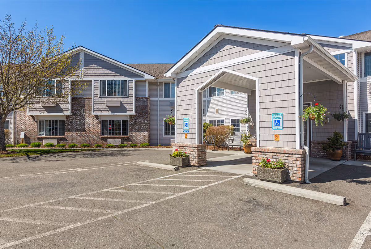 Exterior view of Trustwell Living at Sinclair Place showing a two-story building with a covered entrance, brick and siding facade, handicap parking spaces, and potted plants under a clear blue sky.