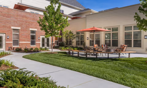 Sunny outdoor courtyard at a senior living facility with patio seating and an umbrella, pathways, lawn, and surrounding building facades.