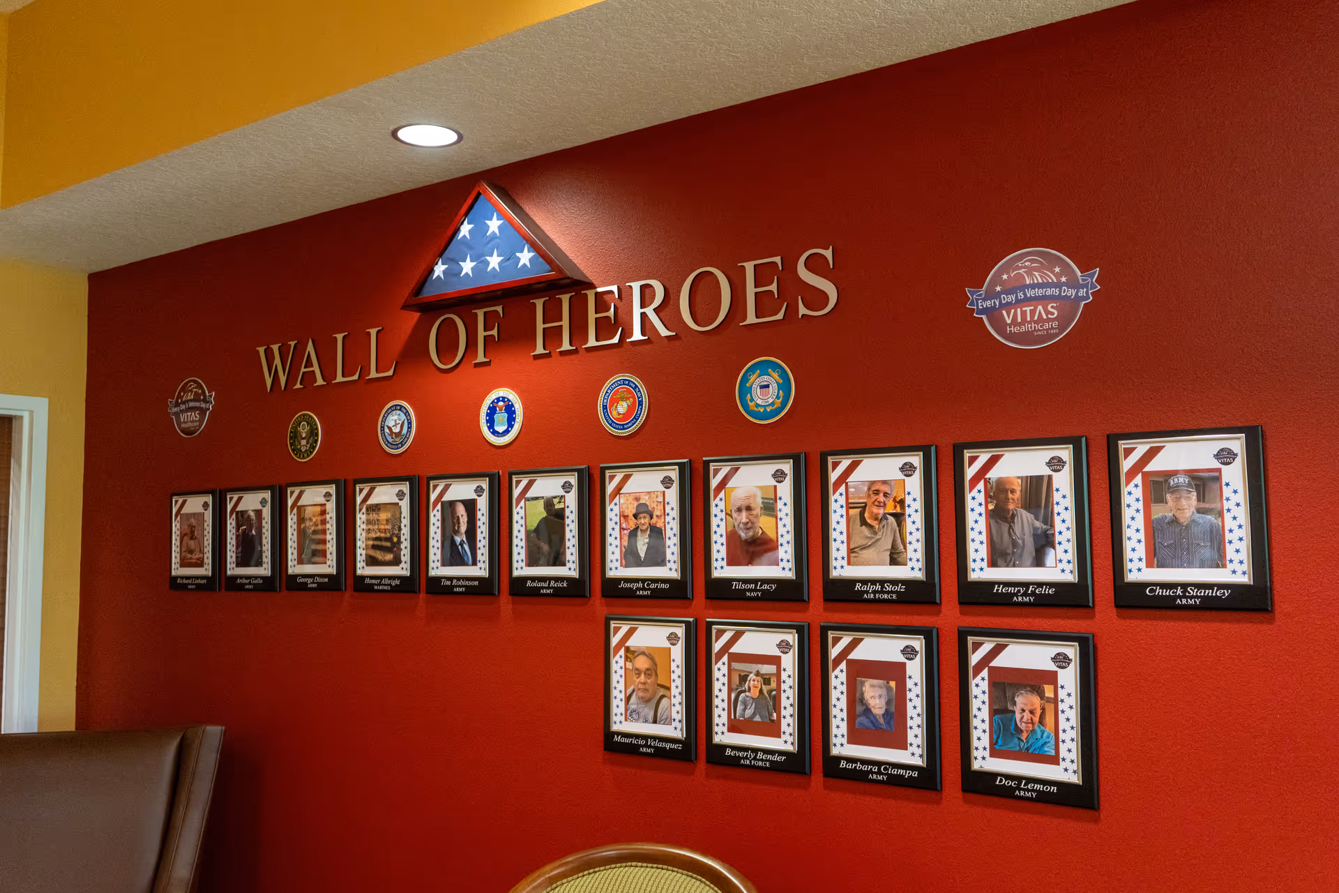 A red wall titled 'Wall of Heroes' displaying framed photos of veterans with their names and military branches beneath each photo. Above the title is a folded American flag in a triangular display case. Various military branch insignias are mounted on the wall, along with VITAS Healthcare logos and slogans.