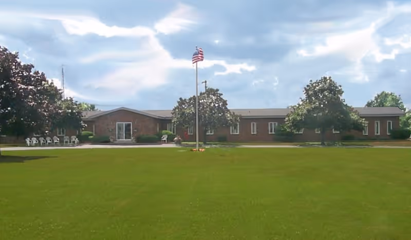 Wide view of a single-story brick building with a large green lawn in front, several trees, and an American flag on a flagpole in the center. White chairs are arranged near the building entrance under a partly cloudy sky.