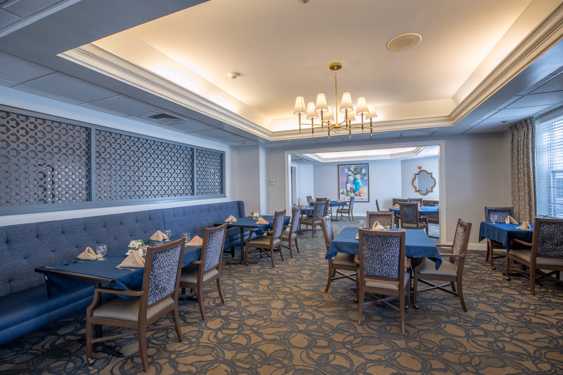Spacious dining room with multiple set tables, upholstered chairs, banquette seating, and a central chandelier.