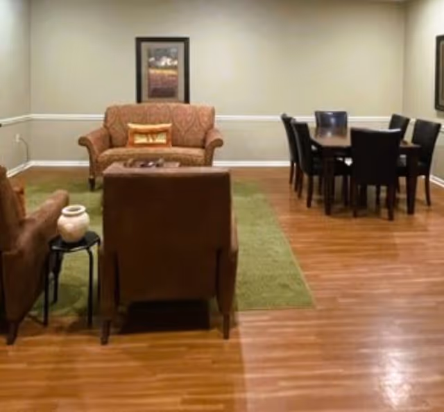 A furnished common room with sofas, armchairs, a green area rug and a dining table with chairs on hardwood floors.