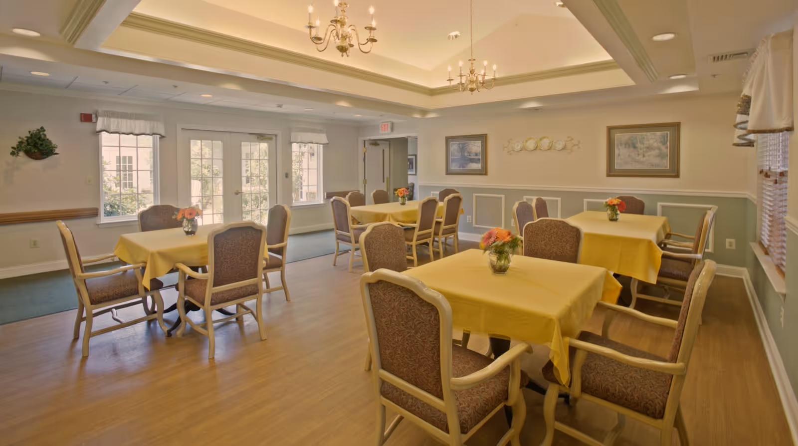 Well-lit dining room with multiple tables draped in yellow tablecloths, upholstered chairs, floral centerpieces, chandeliers, and windows.