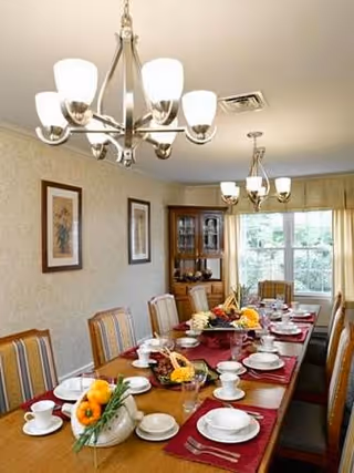 A dining room with a long wooden table set for a meal, featuring white cups, saucers, plates, and silverware on red placemats. The table is decorated with floral centerpieces and a basket of vegetables. The room has striped upholstered chairs, two chandeliers, framed artwork on the walls, a wooden china cabinet, and a large window with a view of greenery outside.