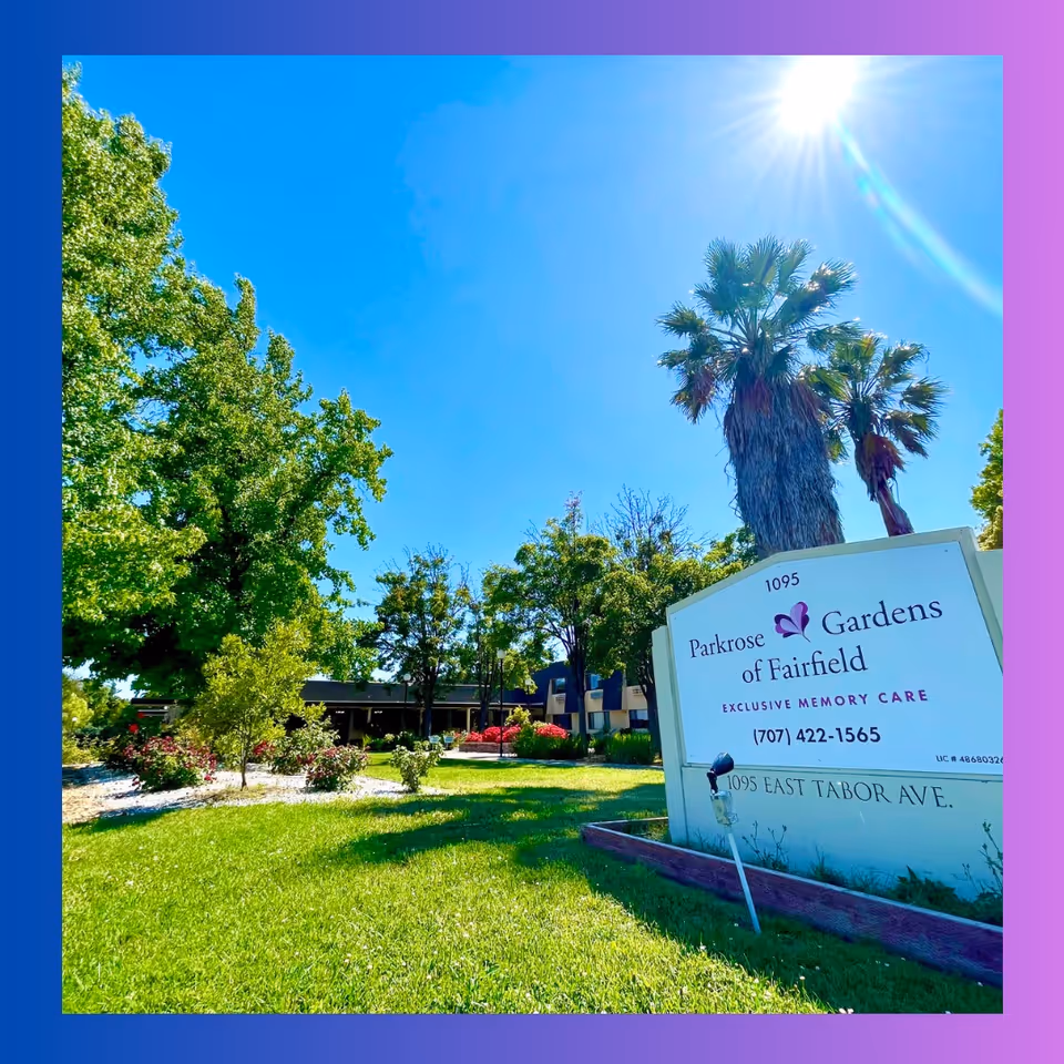 Front lawn and entrance sign for Parkrose Gardens of Fairfield memory care with trees, palm, and the building under a bright blue sky.