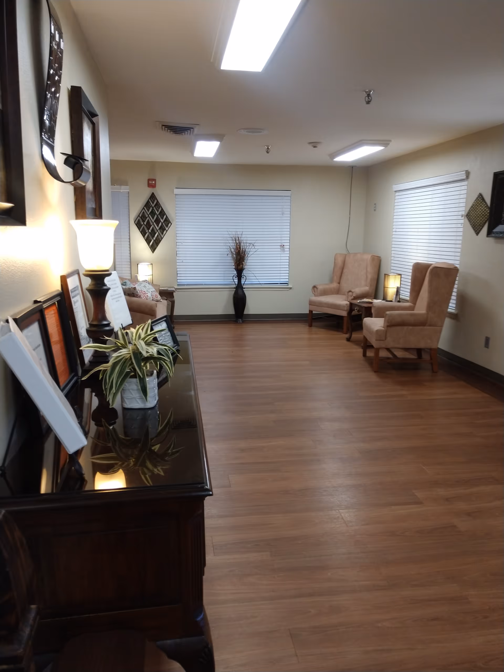 A well-lit room with wooden flooring and beige walls featuring two beige armchairs with a small table and lamp between them near a window with closed blinds. On the left side, there is a dark wooden cabinet with a potted plant, a lamp, and framed pictures or certificates. Decorative wall art is visible on the walls.