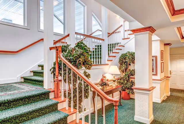 Interior view of a senior living facility featuring a carpeted staircase with green patterned carpet and wooden handrails. There are large windows allowing natural light to brighten the space. A small wooden table with a lamp and floral arrangement is placed against the wall, flanked by two large potted plants. The walls are white with wooden trim, and the hallway extends to a closed door in the background.
