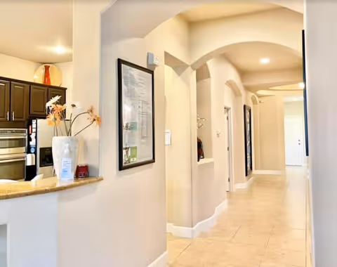 Interior view of a senior care facility hallway with beige walls and tiled floor. On the left side, there is a kitchen area with dark cabinets, a built-in oven, and a countertop with a vase of flowers. The hallway features arched doorways and framed pictures on the walls, leading to a white door at the end.