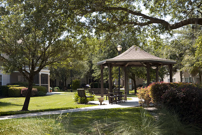 Outdoor garden area at Allegro Senior Living with a wooden gazebo, benches, chairs, and a paved walkway surrounded by green grass, trees, and bushes under a sunny sky.
