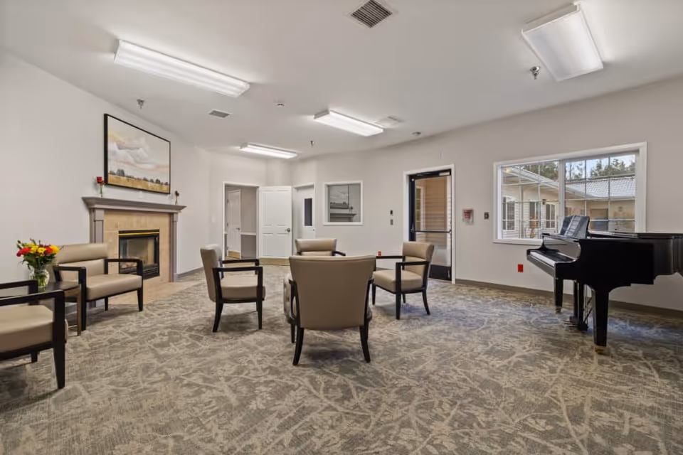 Spacious memory care common room with chairs arranged in a circle, a fireplace, a grand piano, and a large window.