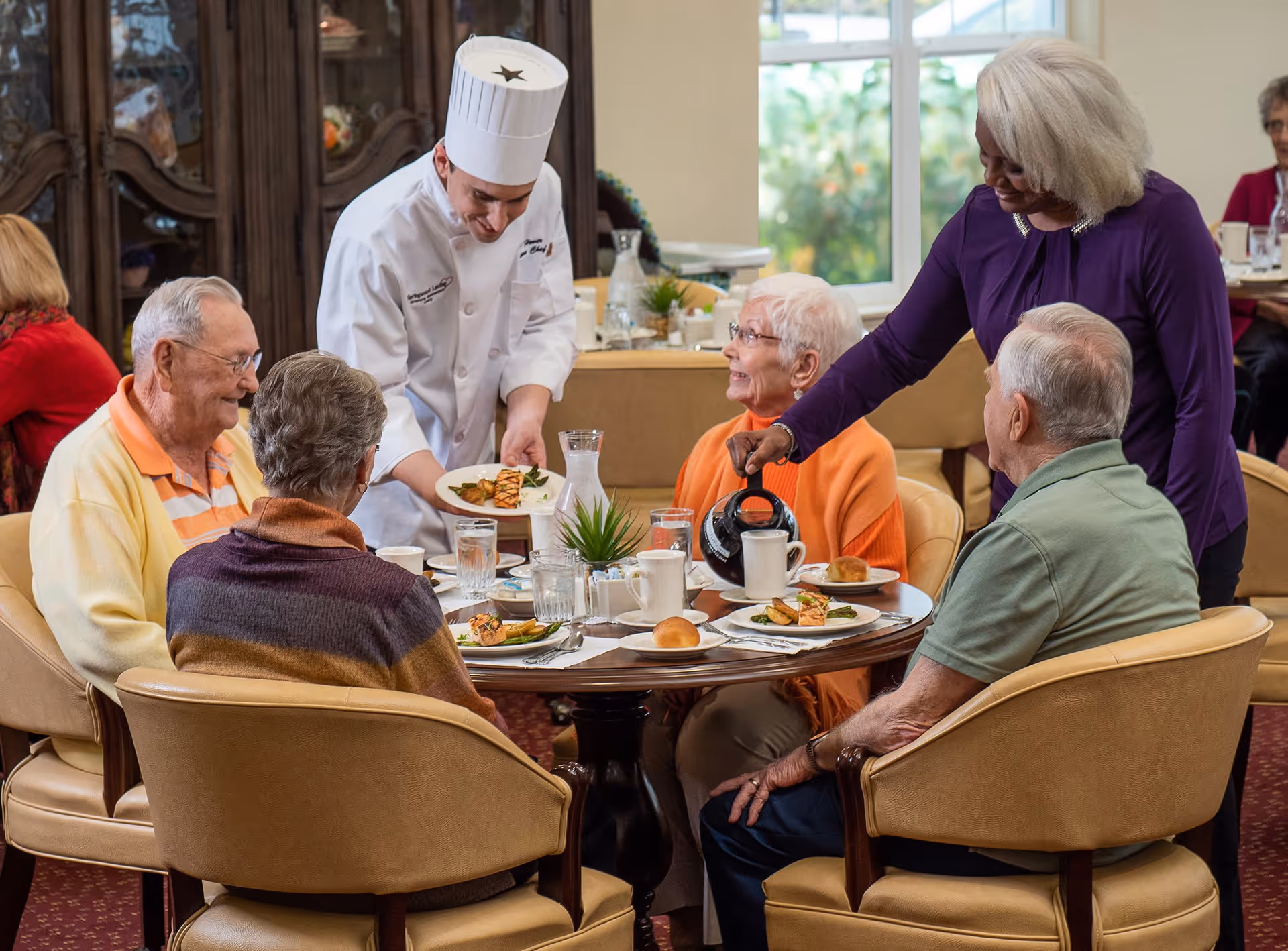 A chef serving plated meals to a group of elderly people seated around a dining table in a well-lit room. A woman is pouring coffee into a cup while the group engages in conversation and smiles. The table is set with plates of food, glasses of water, and a small plant centerpiece.