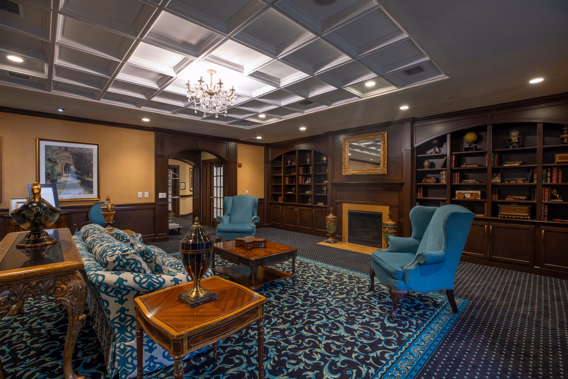 A cozy living room with elegant decor featuring a patterned blue and black carpet, a blue patterned sofa, two blue armchairs, a wooden coffee table, and side tables with decorative vases. The room has dark wood built-in bookshelves filled with books and ornaments, a fireplace with a gold-framed mirror above it, a chandelier hanging from a coffered ceiling, and framed artwork on the wall.