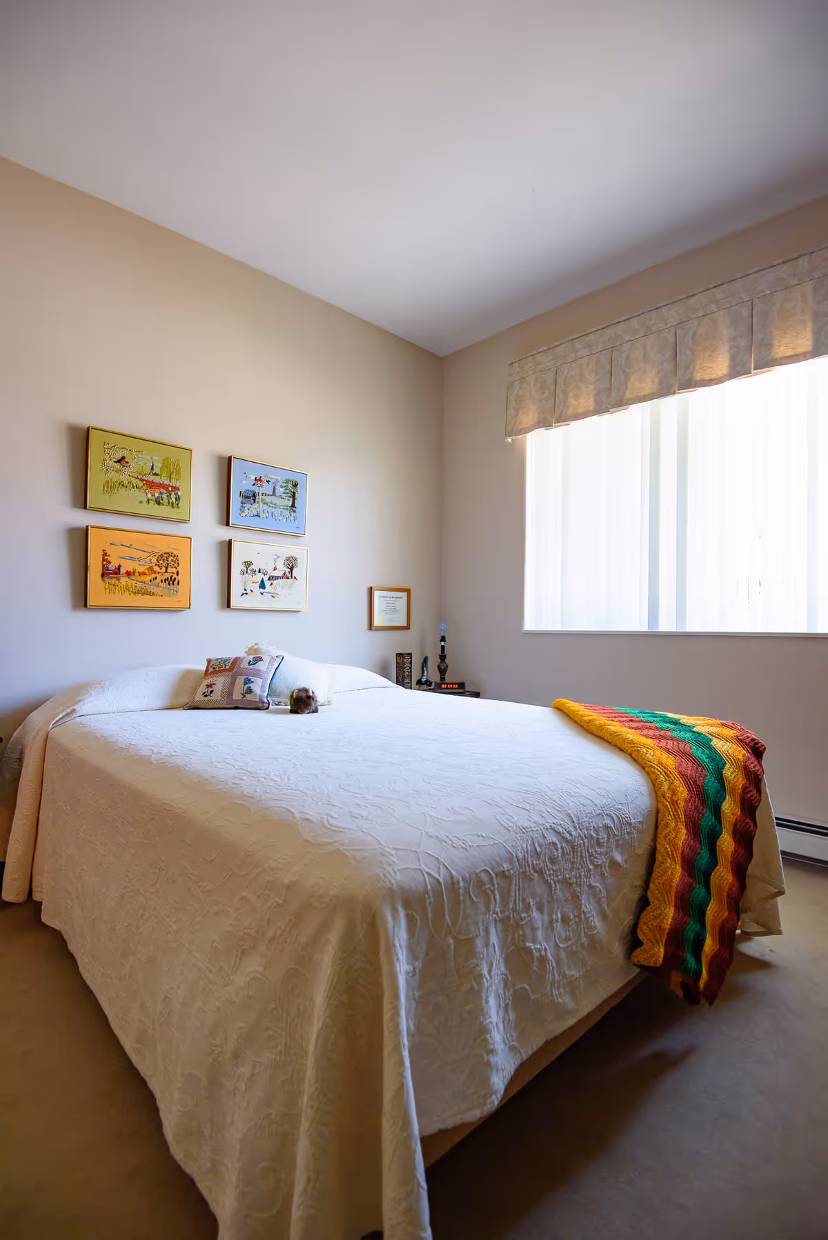 A bedroom with a large bed covered in a white textured bedspread and a colorful knitted blanket draped over the foot. There are two pillows on the bed, one with a floral pattern. Four framed pictures hang on the beige wall above the bed, and a window with vertical blinds and a valance lets in natural light. A small nightstand with a lamp and some decorative items is visible in the corner.