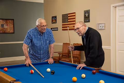 Two elderly men playing pool in a room with a blue pool table. One man is wearing a blue checkered shirt and glasses, while the other is wearing a black sweater and glasses. The room has wall decorations including an American flag, a Marines sign, and a US Army emblem. There are brown chairs against the wall and a closed white door.