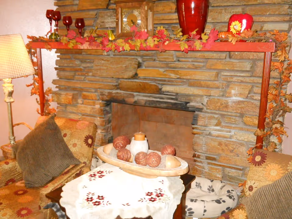 Cozy living room corner featuring a stone fireplace decorated with autumn leaves garland, red wine glasses, a clock, and a red vase on the mantel. In front of the fireplace is a small table with a white embroidered cloth and a decorative bowl with candles and ornamental balls. Two floral-patterned armchairs with brown cushions flank the table, and a floor lamp with a checkered shade stands to the left.