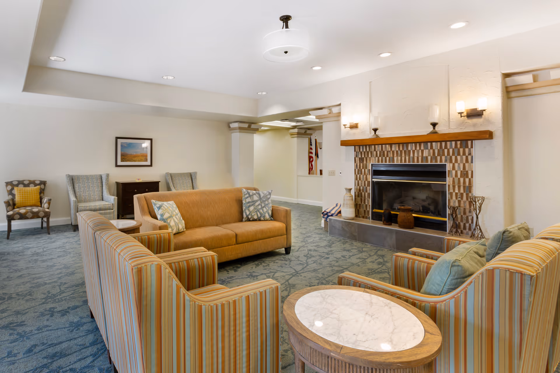 A cozy living room area with a fireplace featuring a patterned tile surround and wooden mantel. The room has a beige sofa, two striped armchairs with green pillows, a small round marble-top table, and additional seating including two patterned armchairs and a small cabinet with a framed picture above it. The carpet is blue with a subtle pattern, and the walls are painted white with recessed lighting and wall sconces providing illumination.