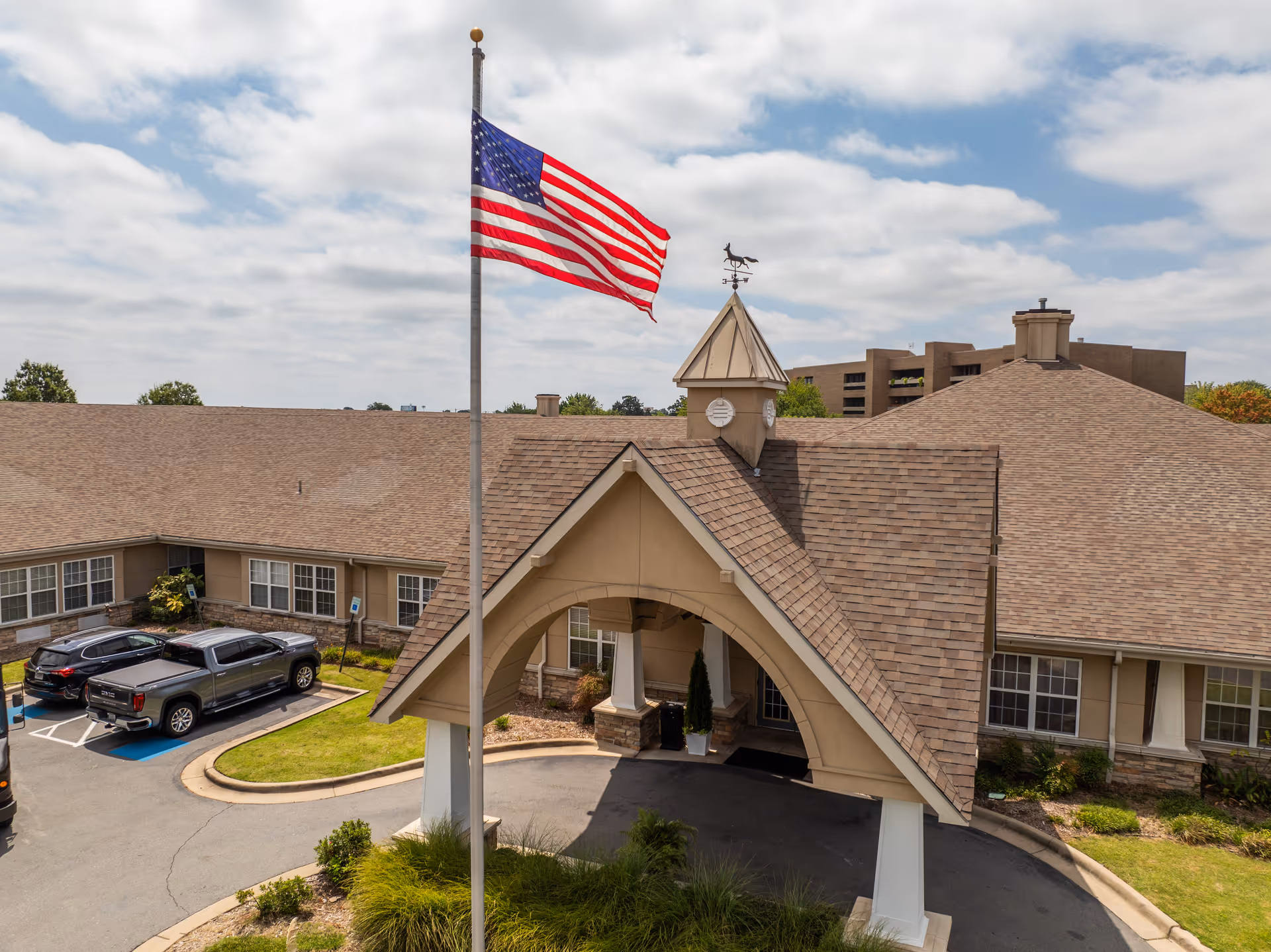 Front entrance of a senior living facility with a covered porte-cochere, parked cars, and an American flag on a tall flagpole.