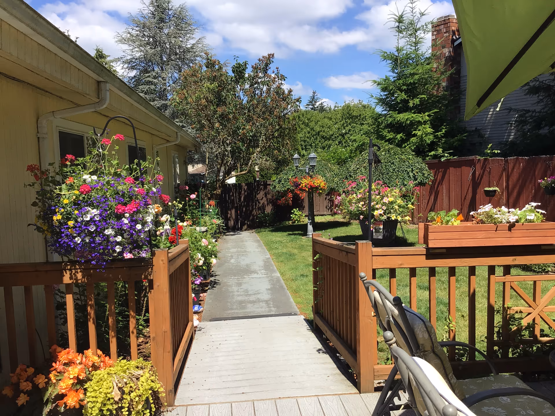 Sunlit backyard pathway between a house and a wooden fence lined with colorful hanging flower baskets and a small deck with chairs.